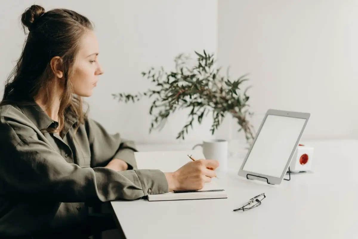 Woman sitting at a white desk writing in a notebook while looking at a tablet screen.
