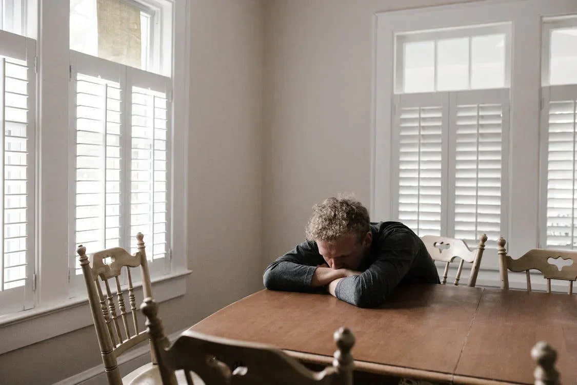 A person resting their head on folded arms at a wooden table in a softly lit room, capturing a quiet moment of overwhelm or emotional exhaustion.