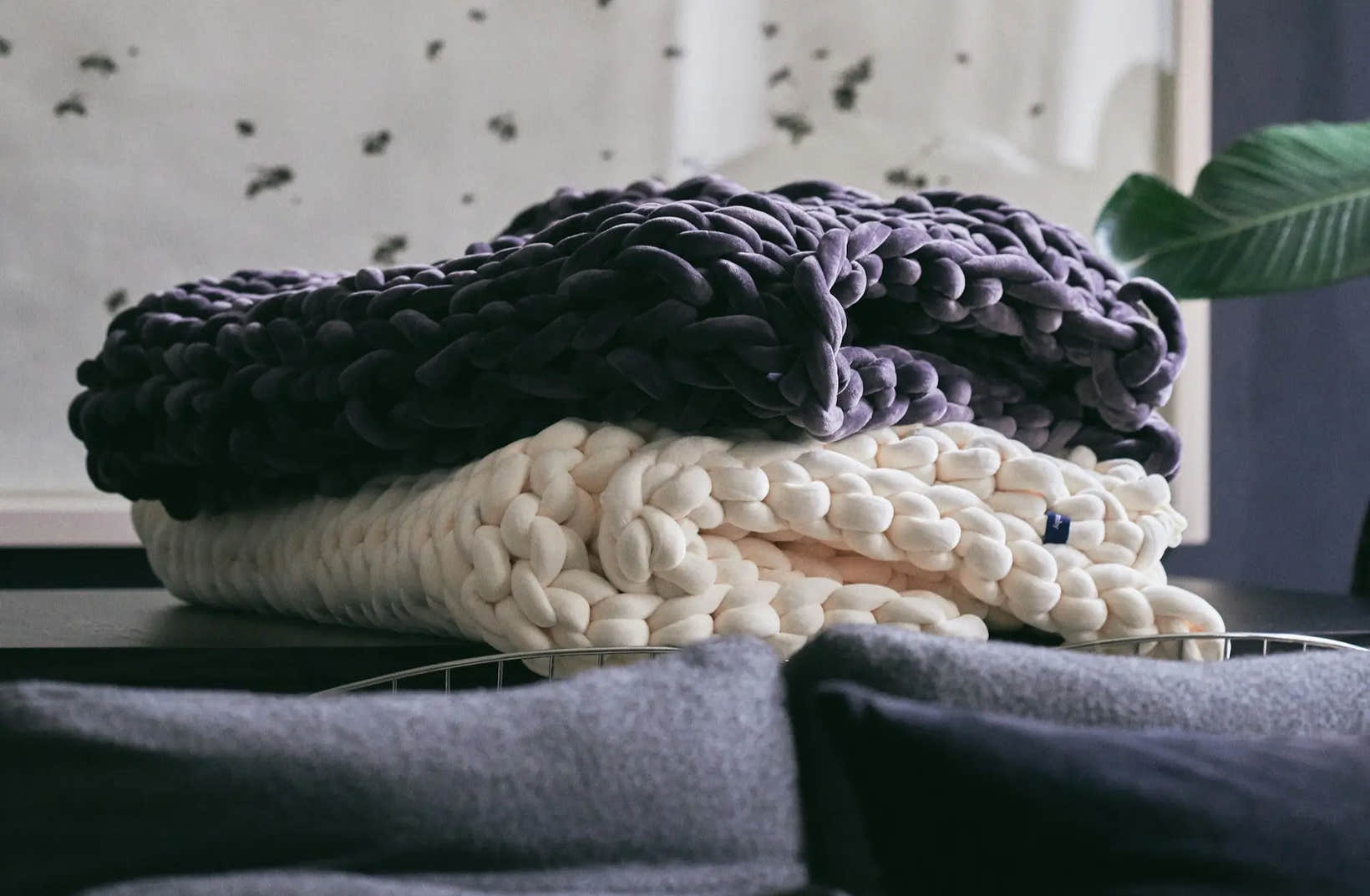 Two chunky knit weighted blankets, one dark purple and one cream, neatly folded and stacked on a table.