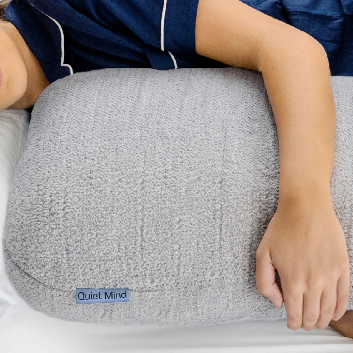 A person lying in bed with their head resting on a large, textured gray pillow. The person is wearing navy blue pajamas with white piping, and the pillow features a small blue label that reads 'Quiet Mind'.