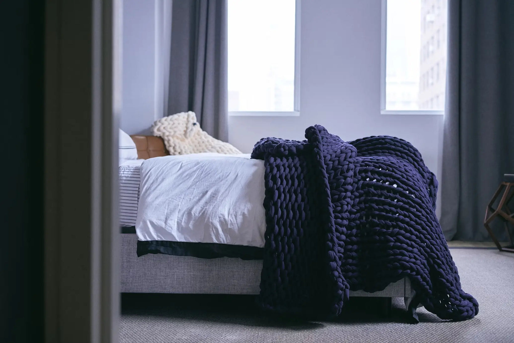 A chunky weighted blanket draped over the edge of a neatly made bed in a softly lit bedroom.
