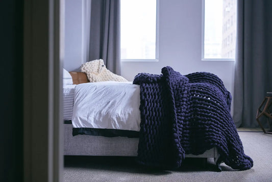 A chunky weighted blanket draped over the edge of a neatly made bed in a softly lit bedroom.