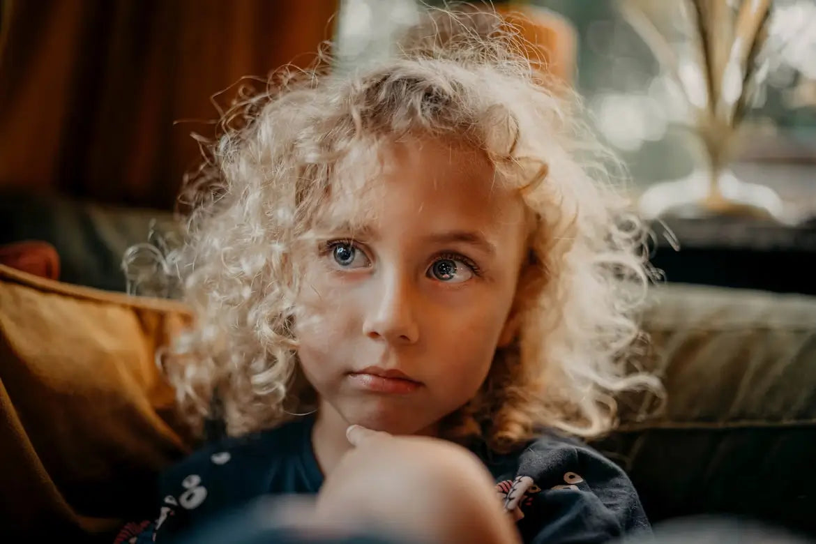 Young child with curly blonde hair and blue eyes sitting indoors, looking thoughtful.