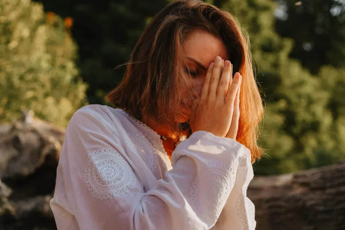 A woman standing outdoors with her hands gently pressed to her face in a calming gesture, symbolizing mindfulness, reflection, and stress relief in a natural setting.