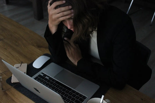 A woman sitting at a desk with her head in her hand, looking stressed while working on a laptop—illustrating the overwhelm that often comes with overthinking and chronic stress.