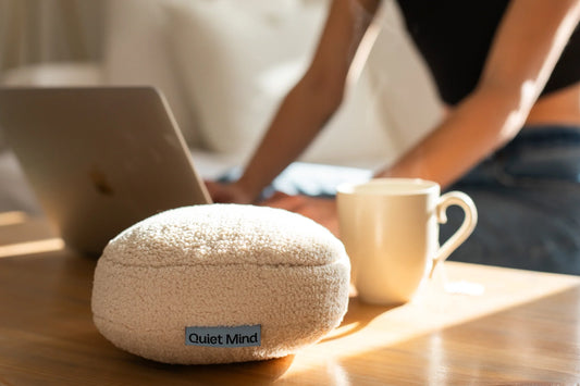 Cream Quiet Mind weighted stress ball sits beside a laptop and mug on a sunlit desk.