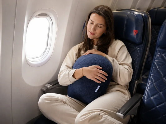 Woman relaxes on a plane while hugging a navy Quiet Mind weighted pillow.