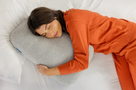 Woman rests on a bed while hugging a gray Quiet Mind weighted body pillow.