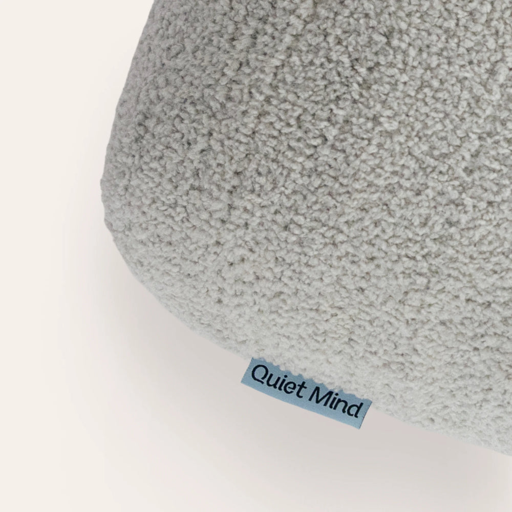 Close-up of a light gray textured plush cushion with a small blue tag reading 'Quiet Mind' on a white background.
